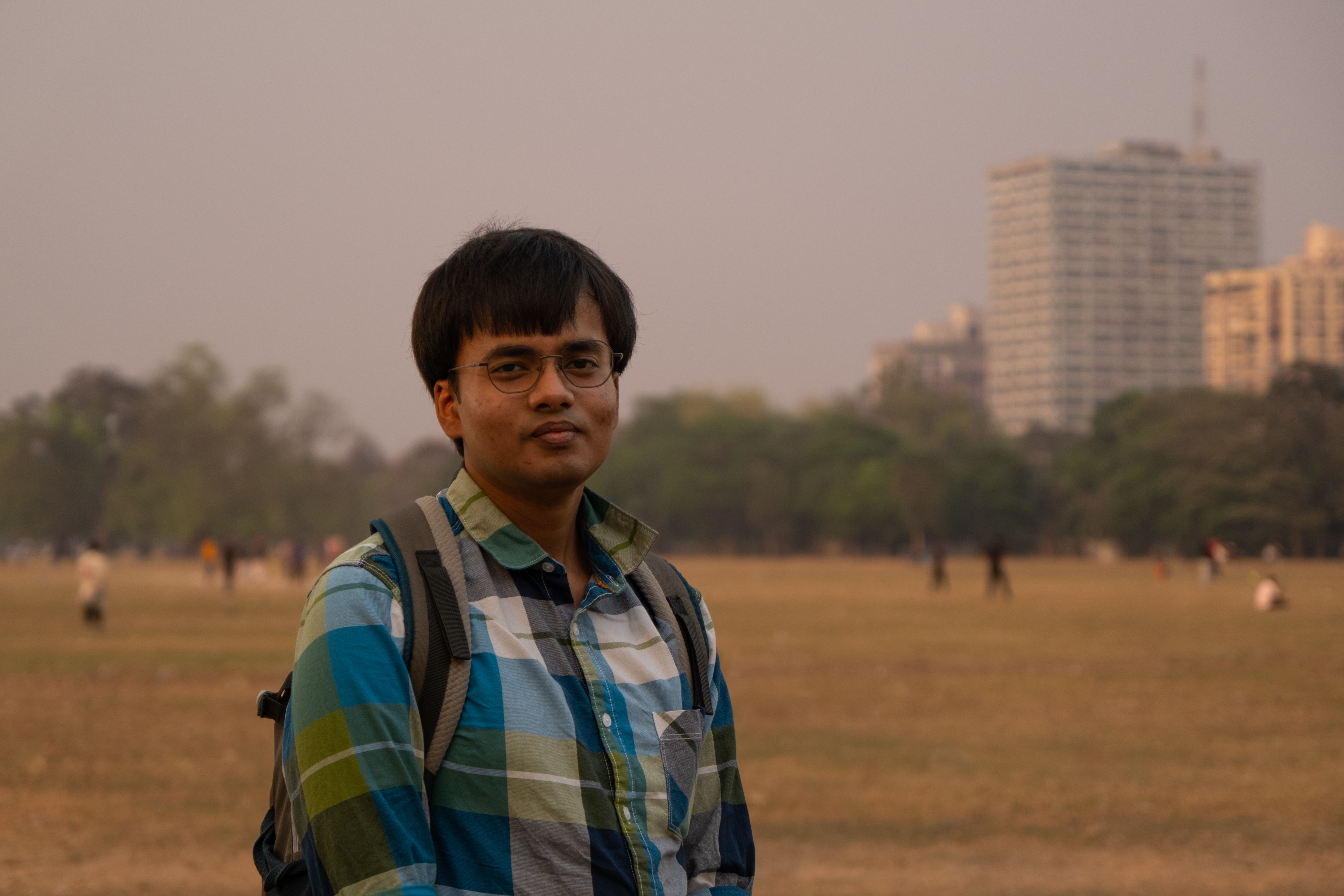 A young man with a backpack stands in a park with a city skyline in the background.