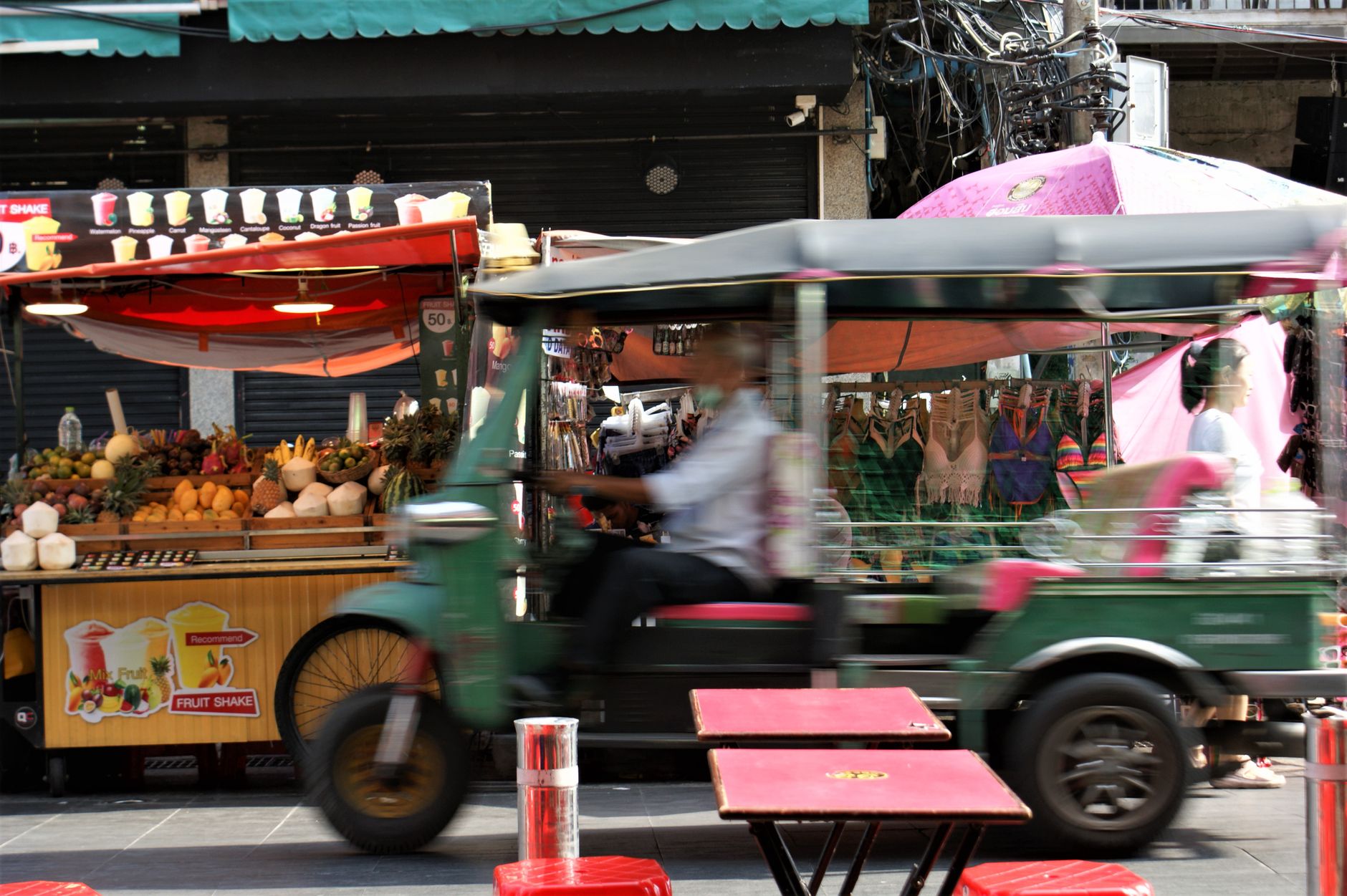 Vibrant street scene in Bangkok showing a moving tuk-tuk blurring past colourful market food stalls.