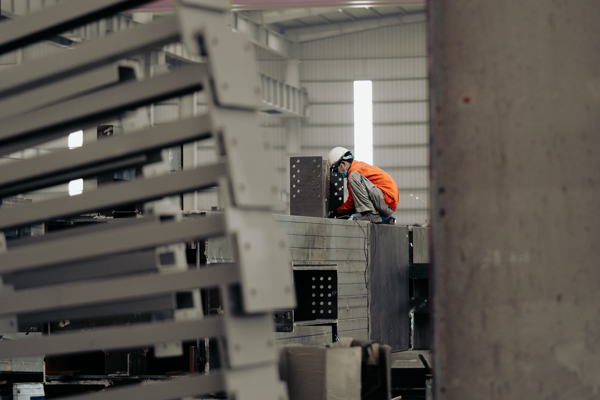 Worker in orange safety suit and white helmet inspecting metal structures in a factory.