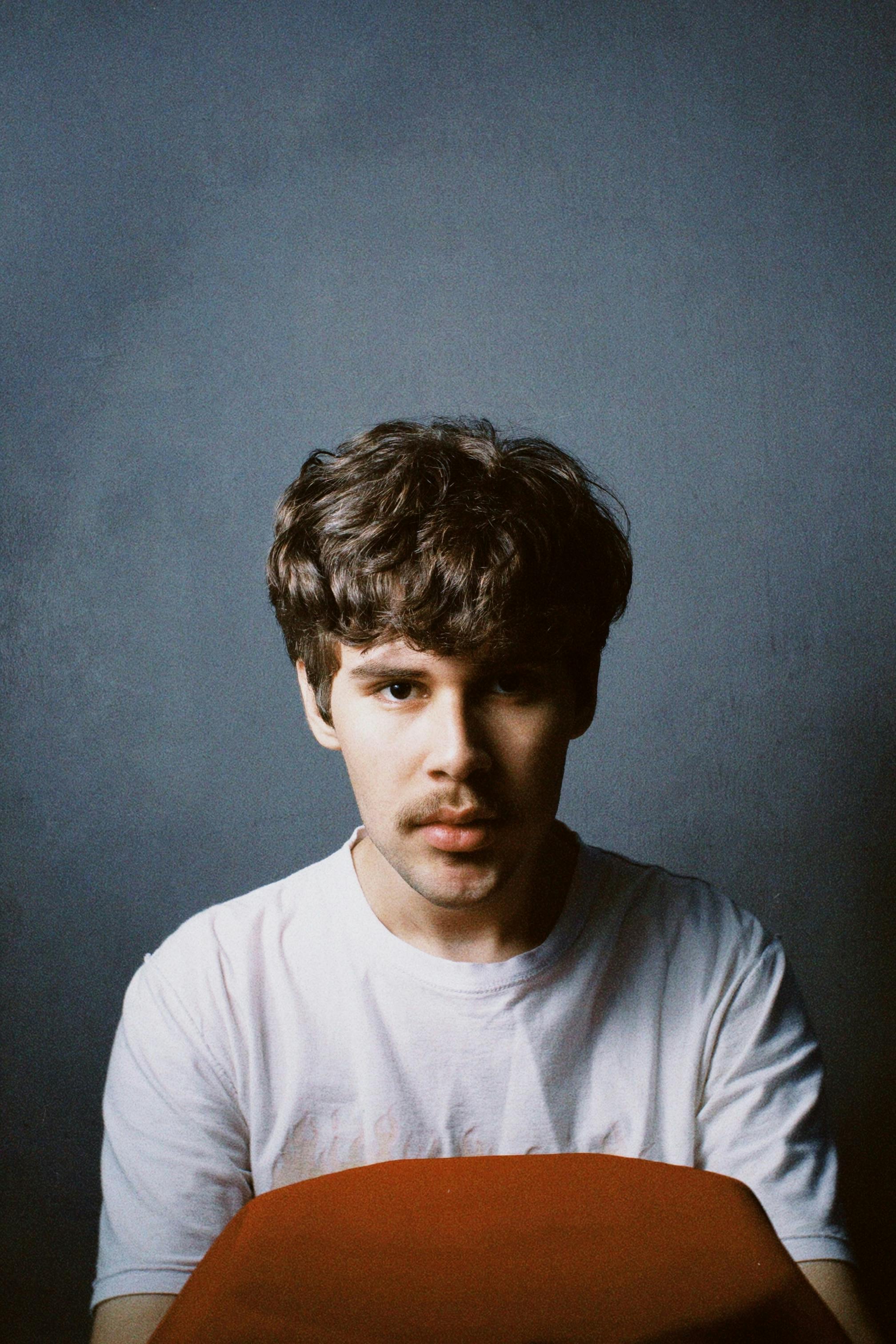 Studio portrait of a young man with a mustache wearing a white t-shirt against a textured background.