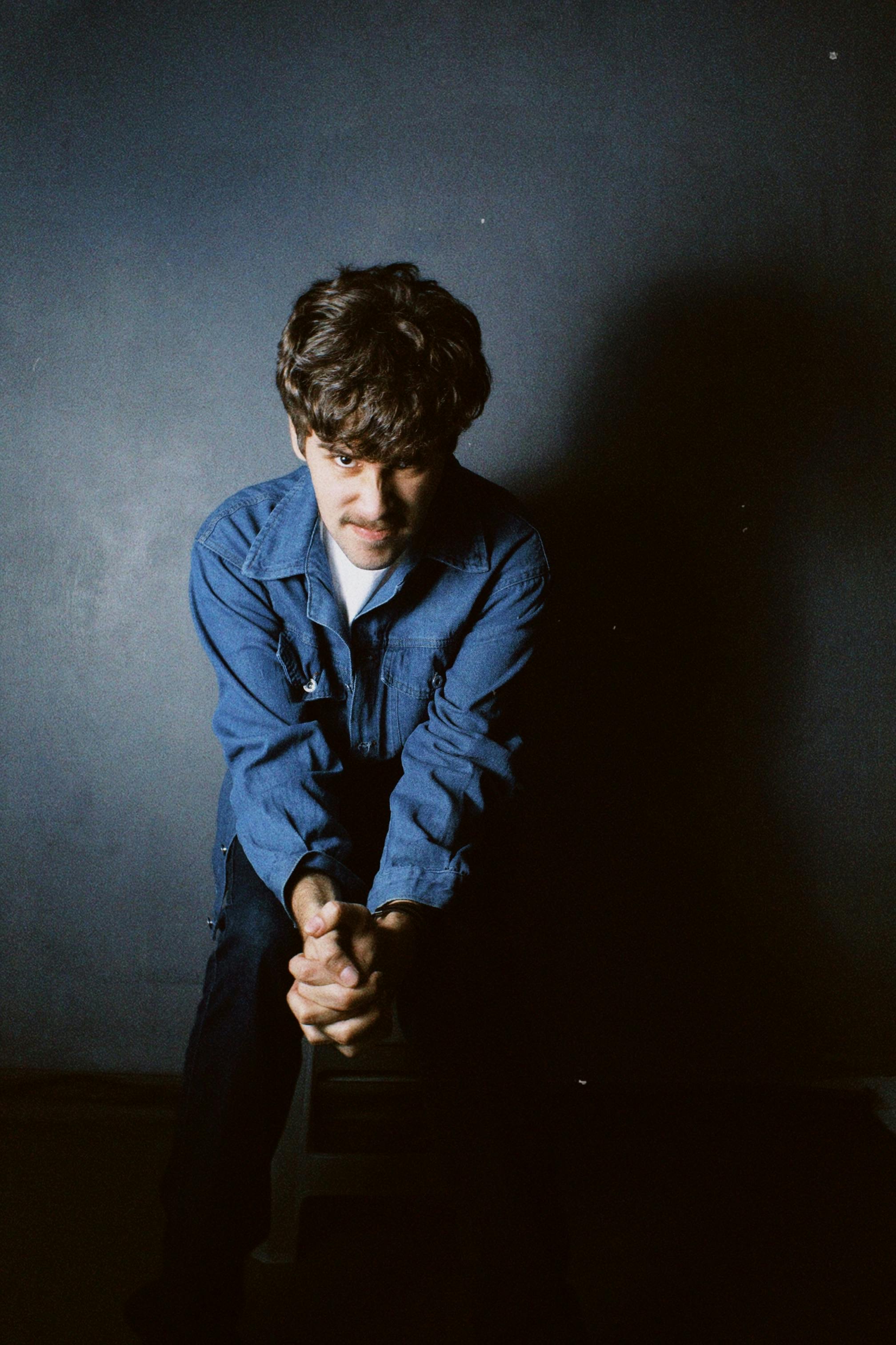 A contemplative man in a denim shirt sits against a dark background, evoking a moody tone.