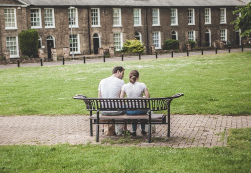 Free A couple enjoys a peaceful moment on a park bench with historic brick buildings in the background. Stock Photo