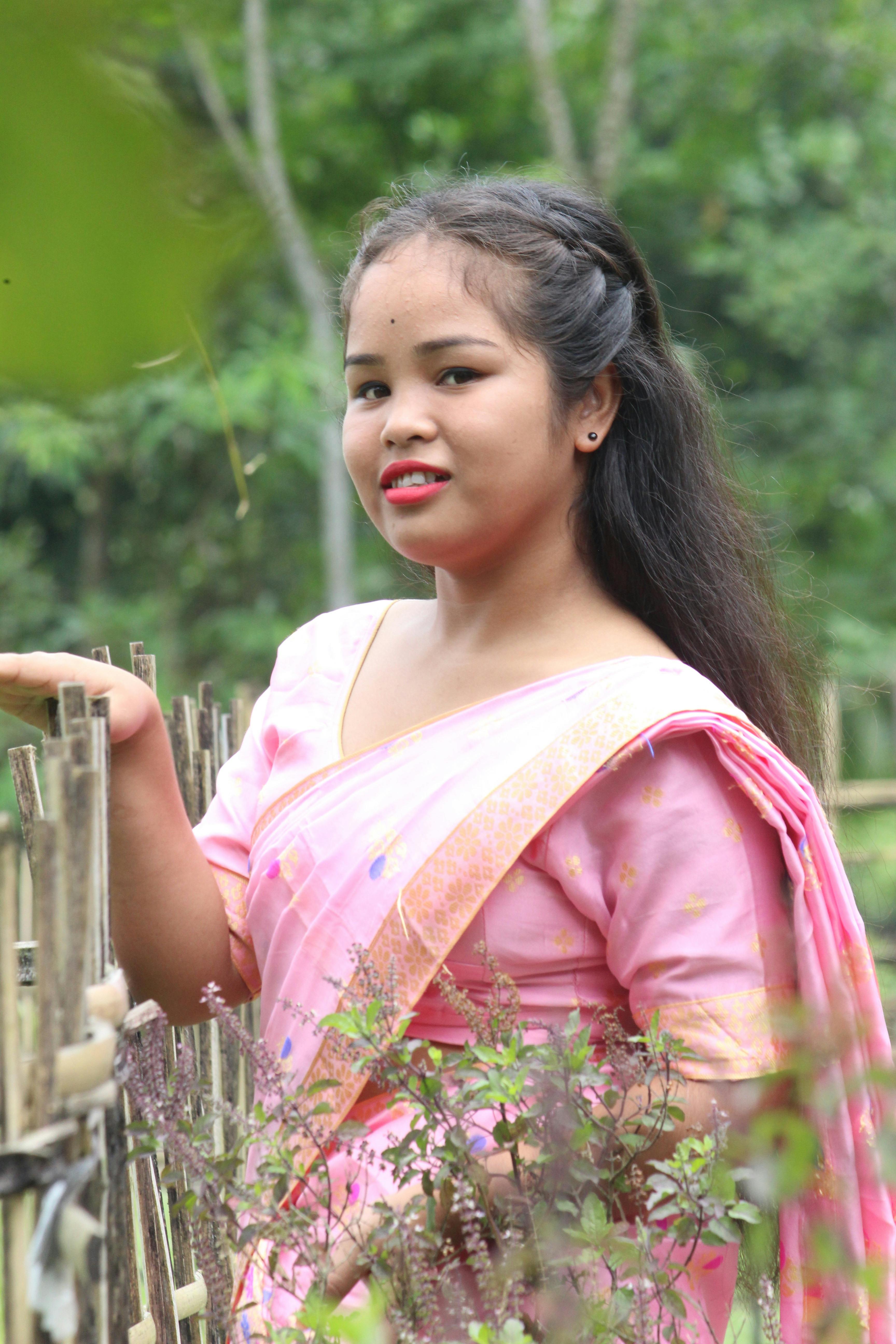 Asian woman in traditional pink saree leaning against a rustic fence outdoors.