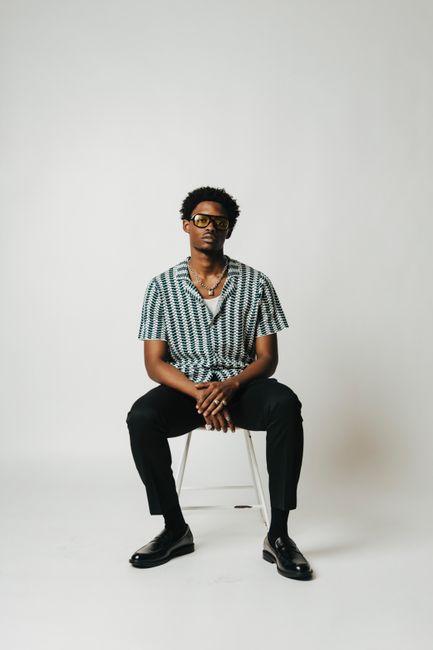 A fashionable man in eyeglasses and patterned shirt sitting on a chair in a studio setting.