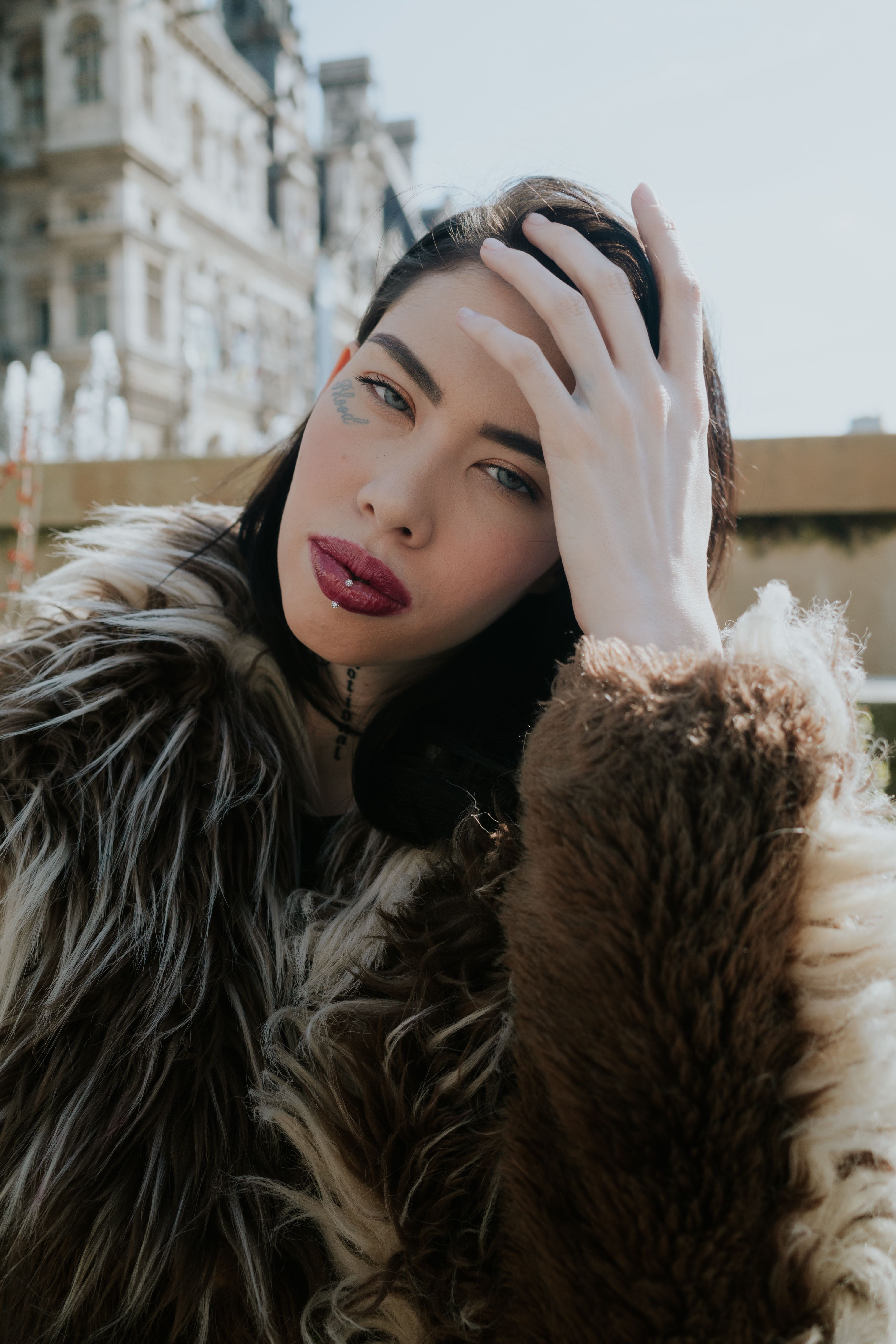 Stylish woman in fur coat posing outdoors with hand in hair, striking red lips.