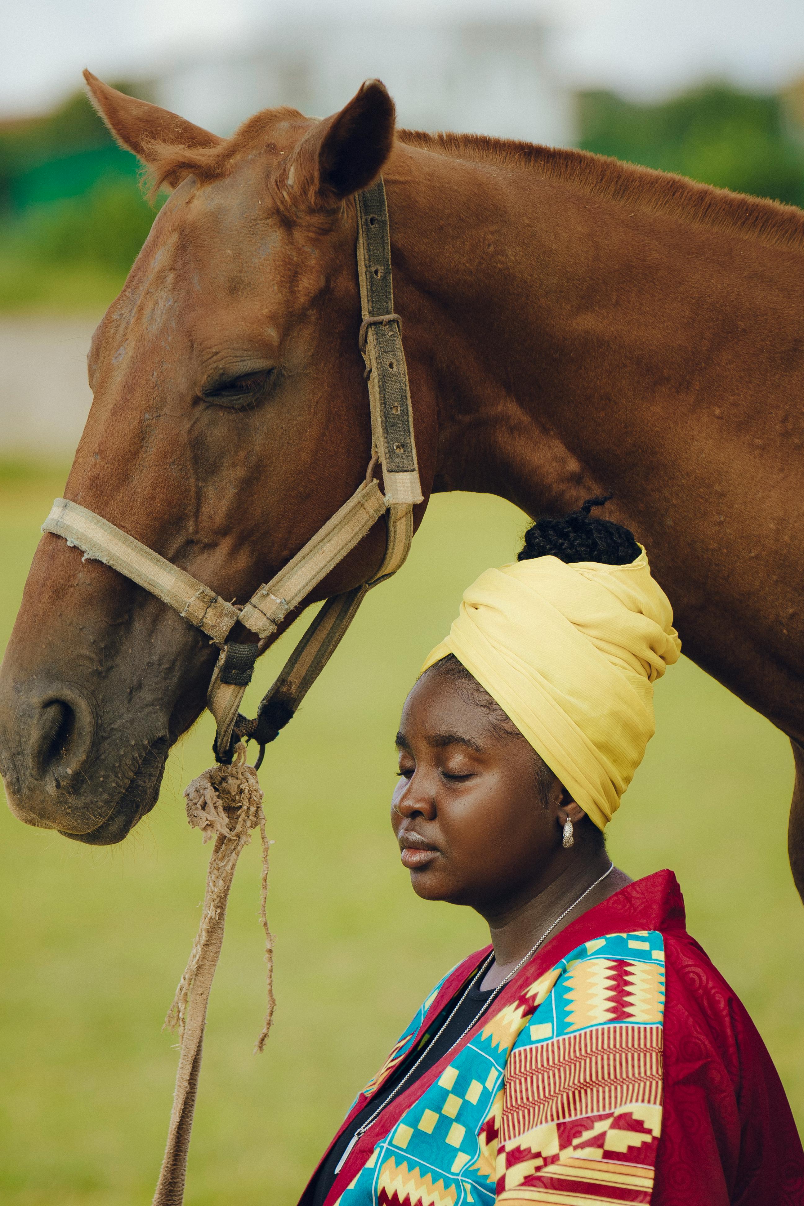 Portrait of an African woman in colorful attire standing beside a brown horse in the countryside.