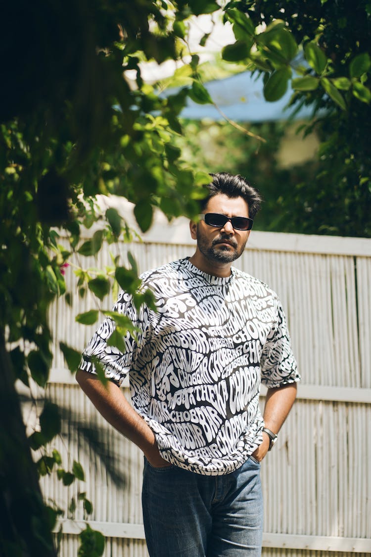 A Man In A Casual Outfit And Sunglasses Standing In The Garden 