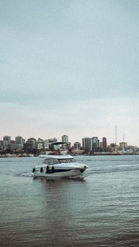 A sleek motorboat gliding in front of a city's coastal skyline under a cloudy sky.