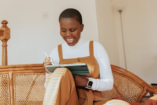 A cheerful woman in overalls sits on a rattan sofa using a tablet. Bright indoor setting.