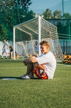 A young man wearing sandals sits and smiles on a sunny soccer field.