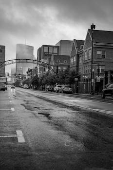 Black and white photo of a wet city street with historic buildings and modern skyline in a rainy urban district.
