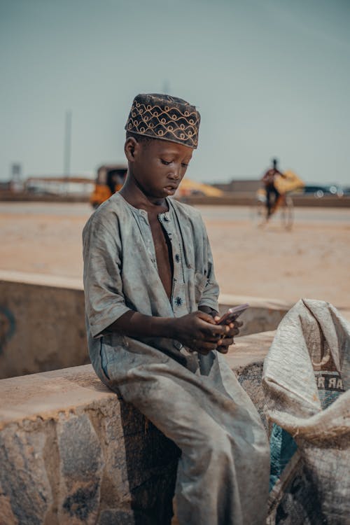 A Boy Wearing a Traditional Gown and Cap Sitting Outside and Using a Smartphone 