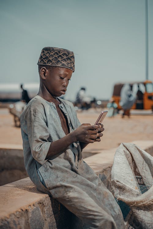 A Boy Wearing a Traditional Gown and Cap Sitting Outside and Using a Smartphone