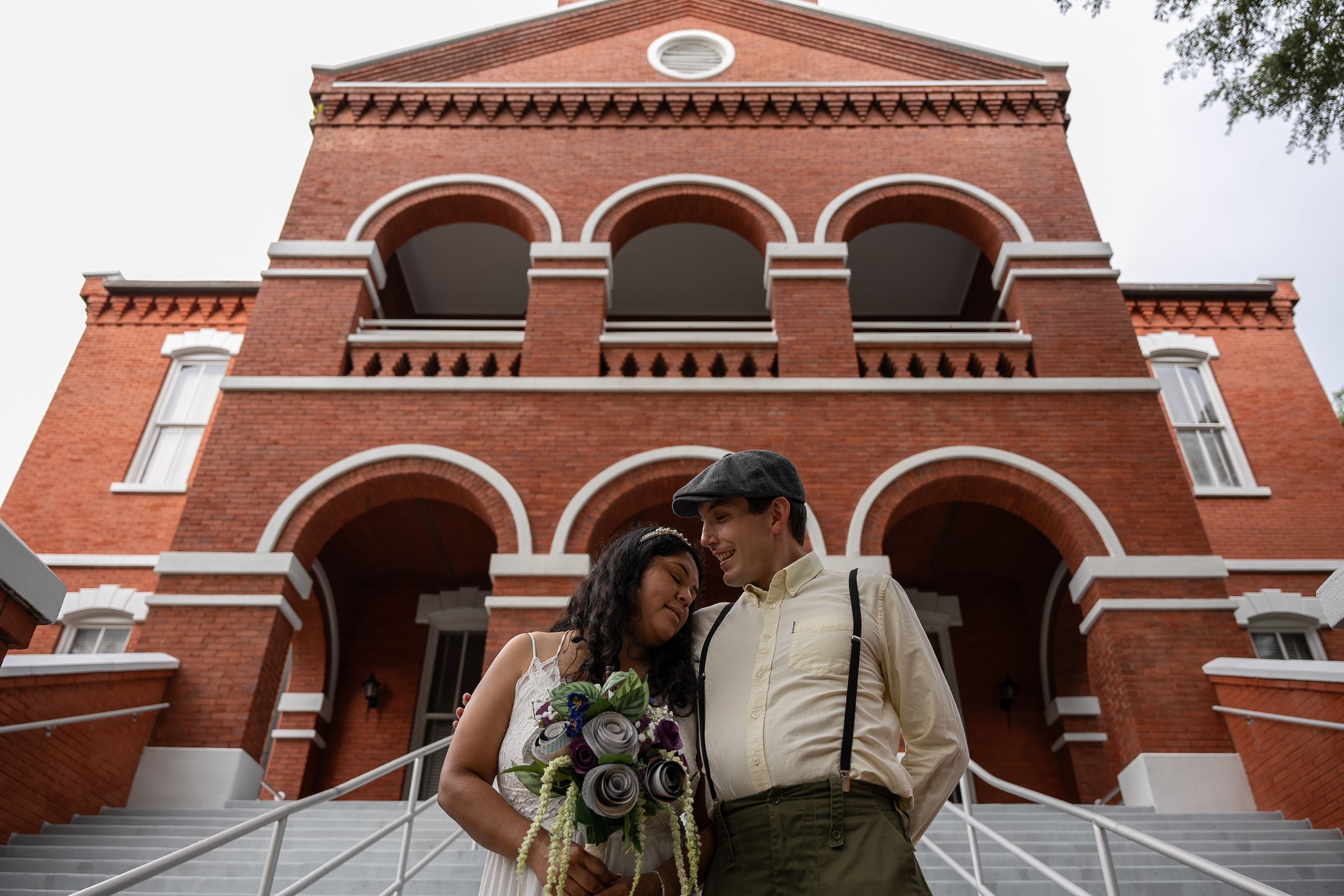 A joyful couple poses with a bouquet on the steps of a historic red brick building.