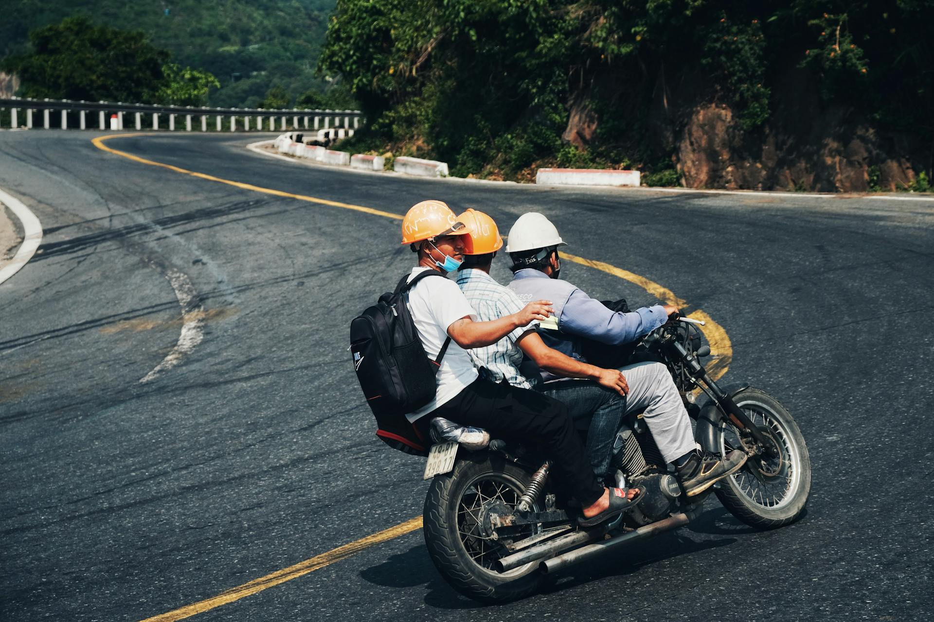 Three men with hard hats ride a motorcycle on a curved mountain road, emphasizing risk and adventure.