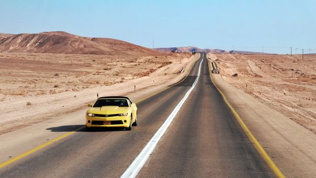 A yellow sports car travels on an empty desert highway under a clear blue sky.