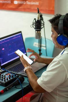 Person managing audio production using a laptop, microphone, and smartphone.