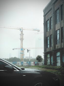 A foggy cityscape featuring construction cranes among buildings and a car nearby, creating a moody urban scene.