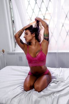 Sensual portrait of a woman in pink lace lingerie with tattoos, striking a pose on a bed.