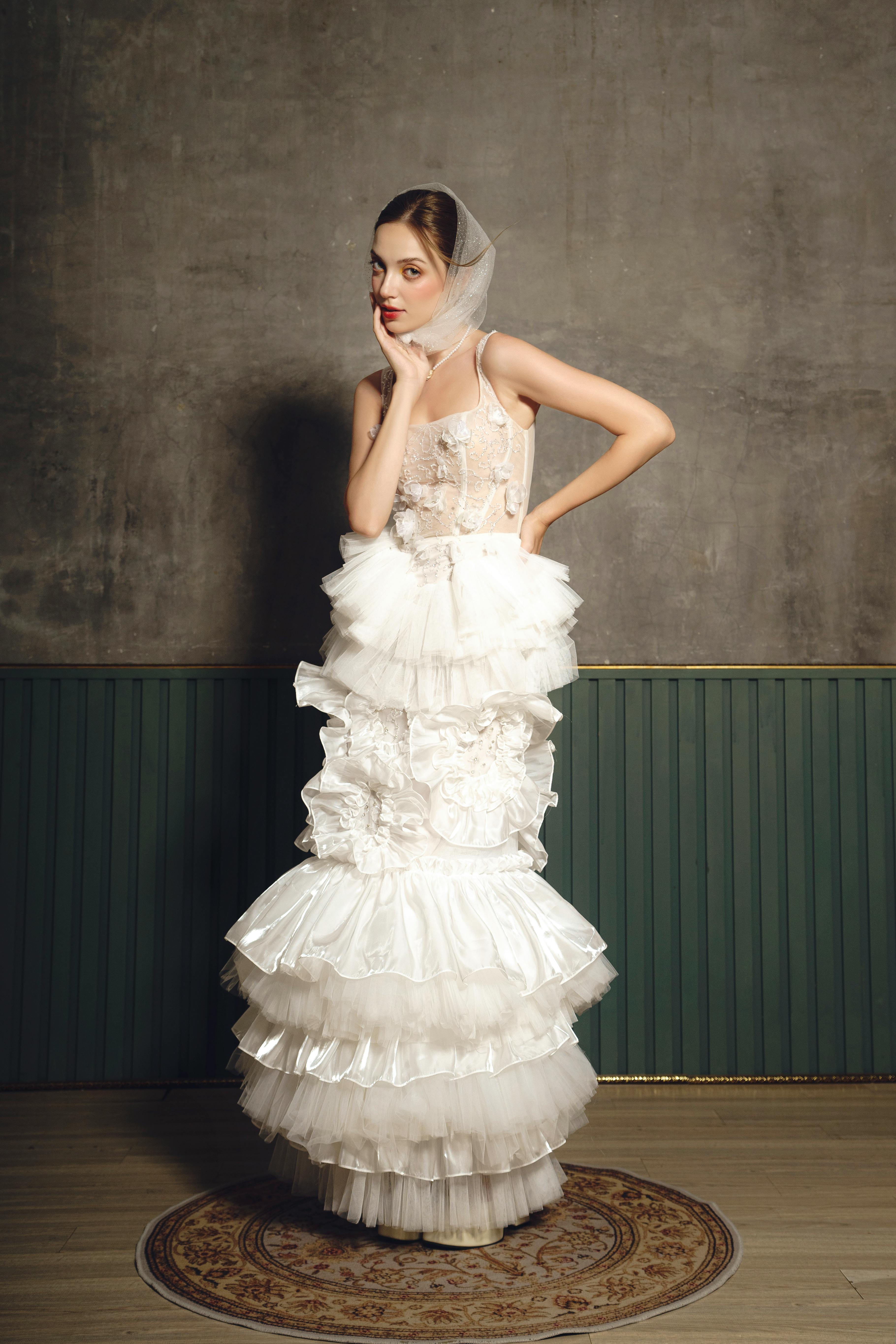 A fashionable woman in a layered white dress poses elegantly indoors, showcasing evening wear.