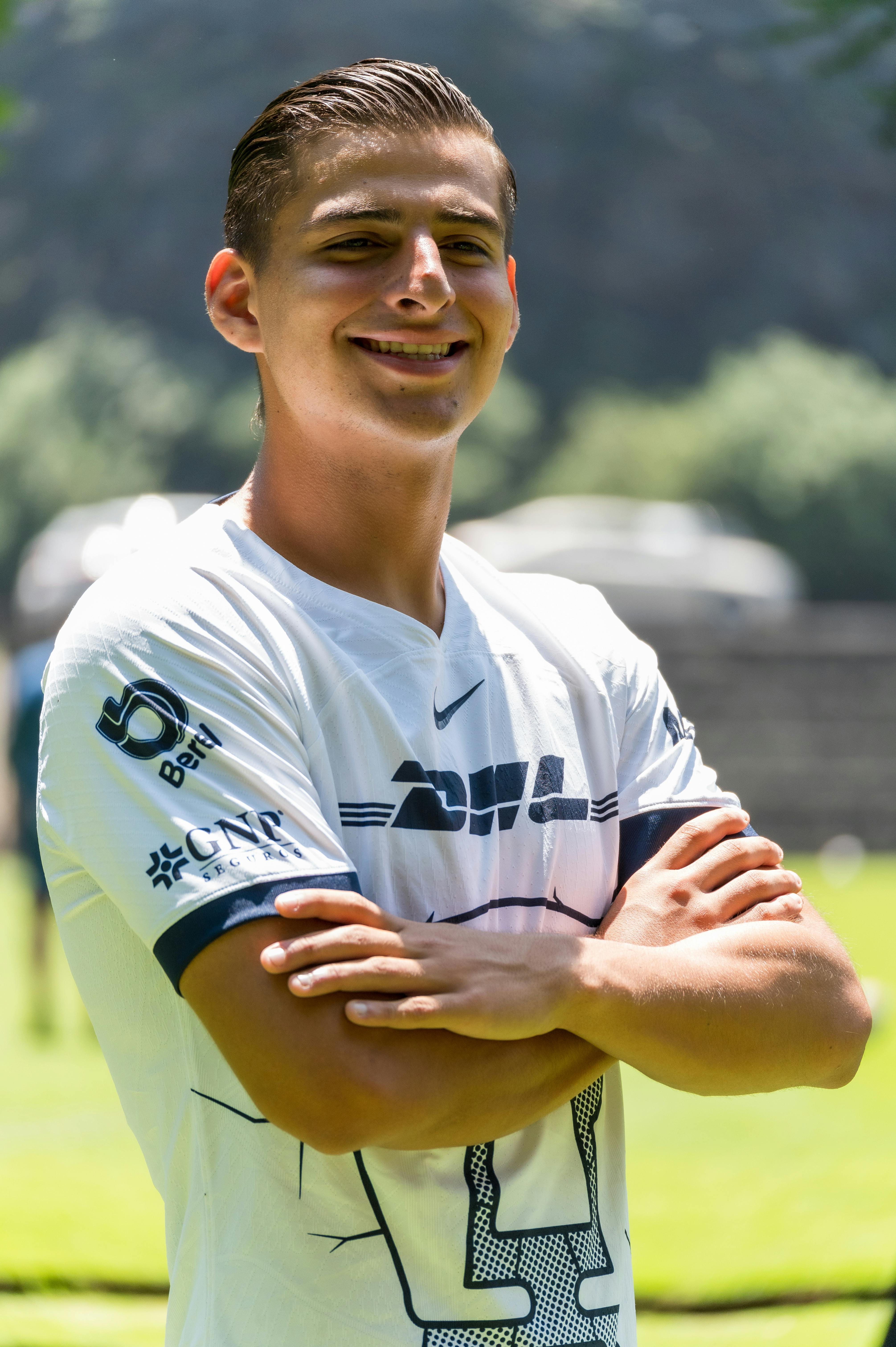 Confident young man smiling with arms crossed wearing a sports jersey outdoors in sunny weather.