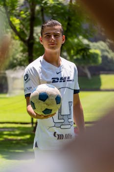 A young male footballer wearing a sports jersey holds a soccer ball in a sunny park.