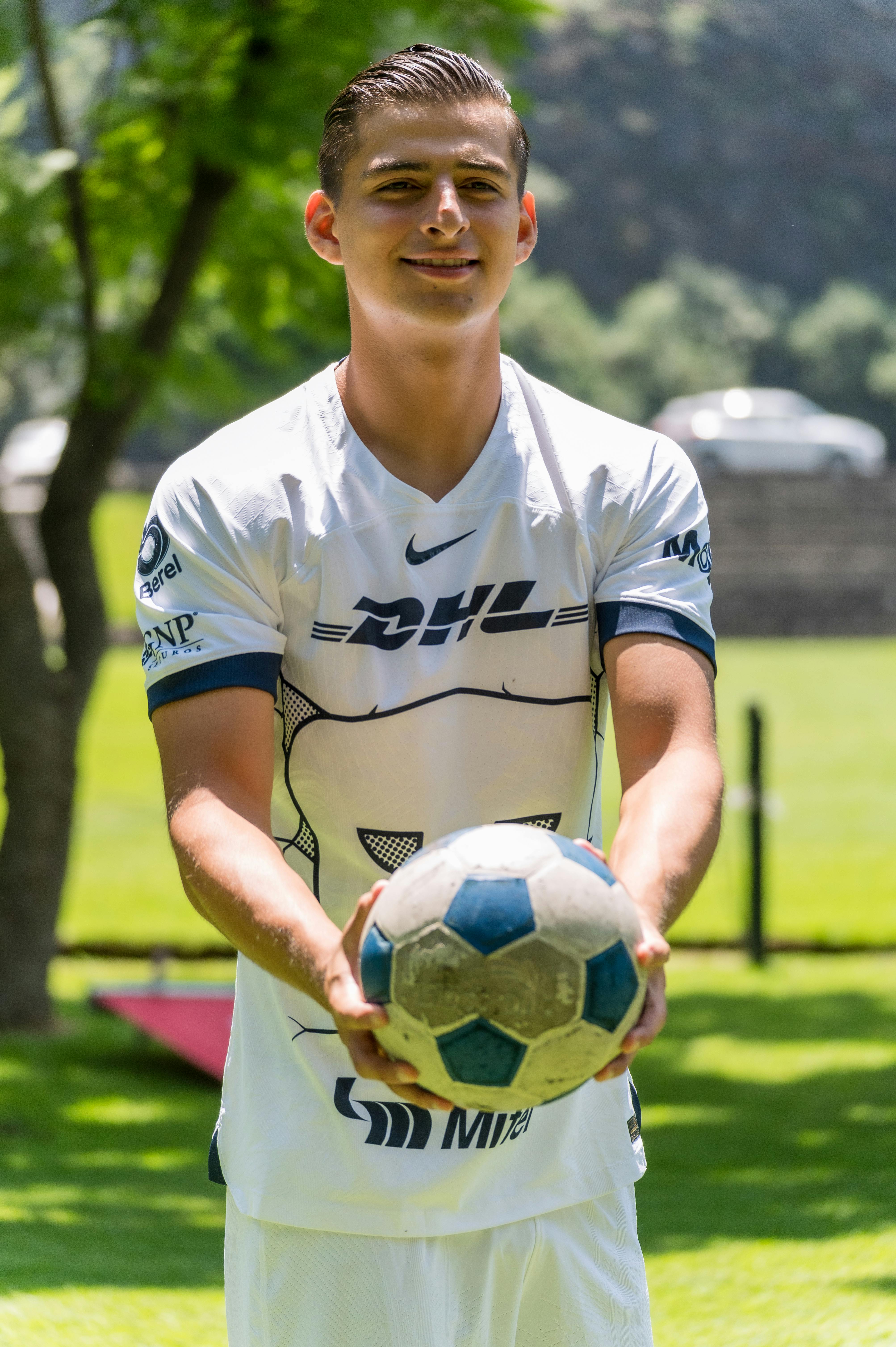 A young male soccer player in a white jersey holds a football outdoors on a sunny day.