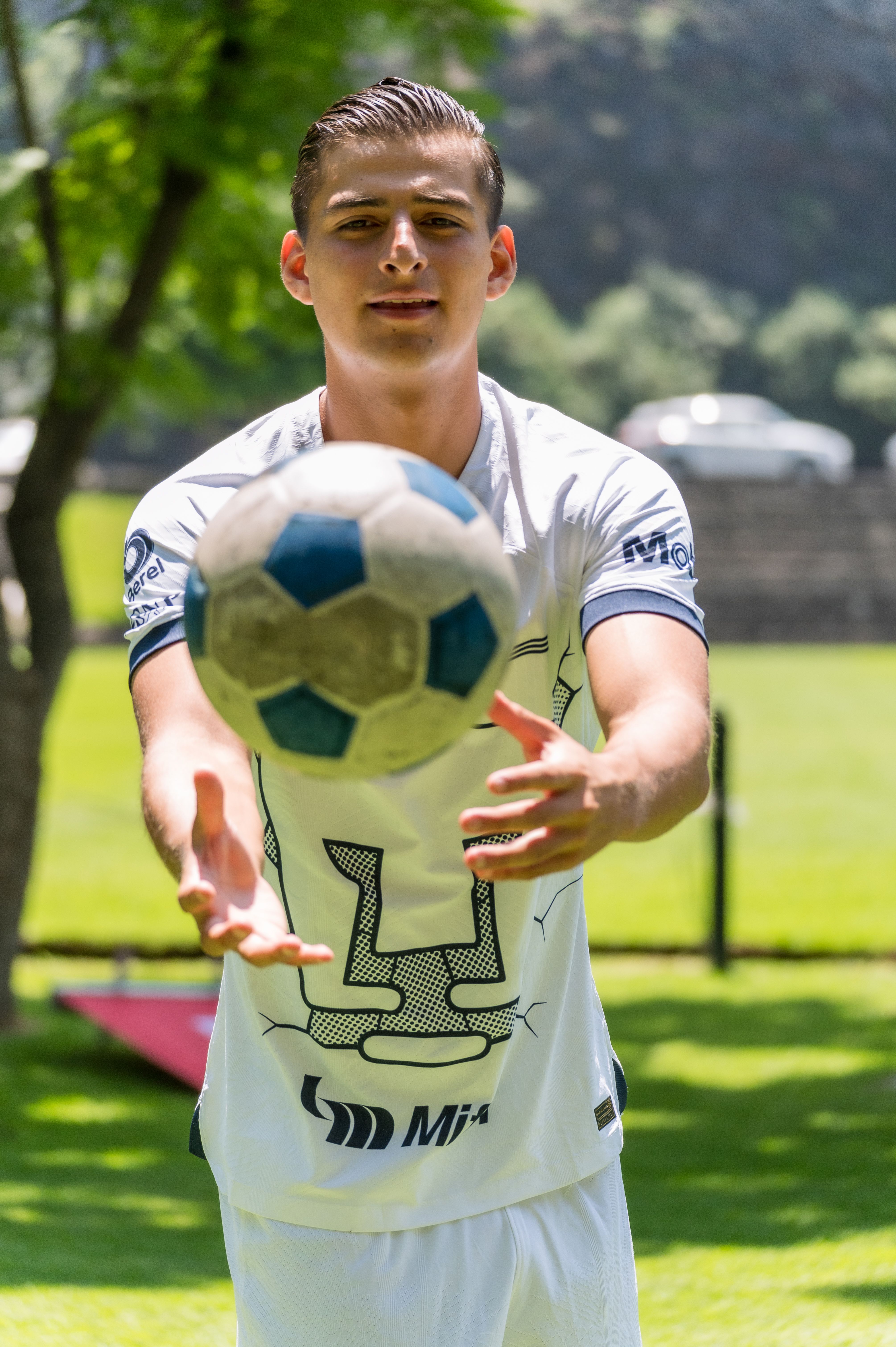 A young man in sportswear catching a soccer ball outdoors in a park on a sunny day.