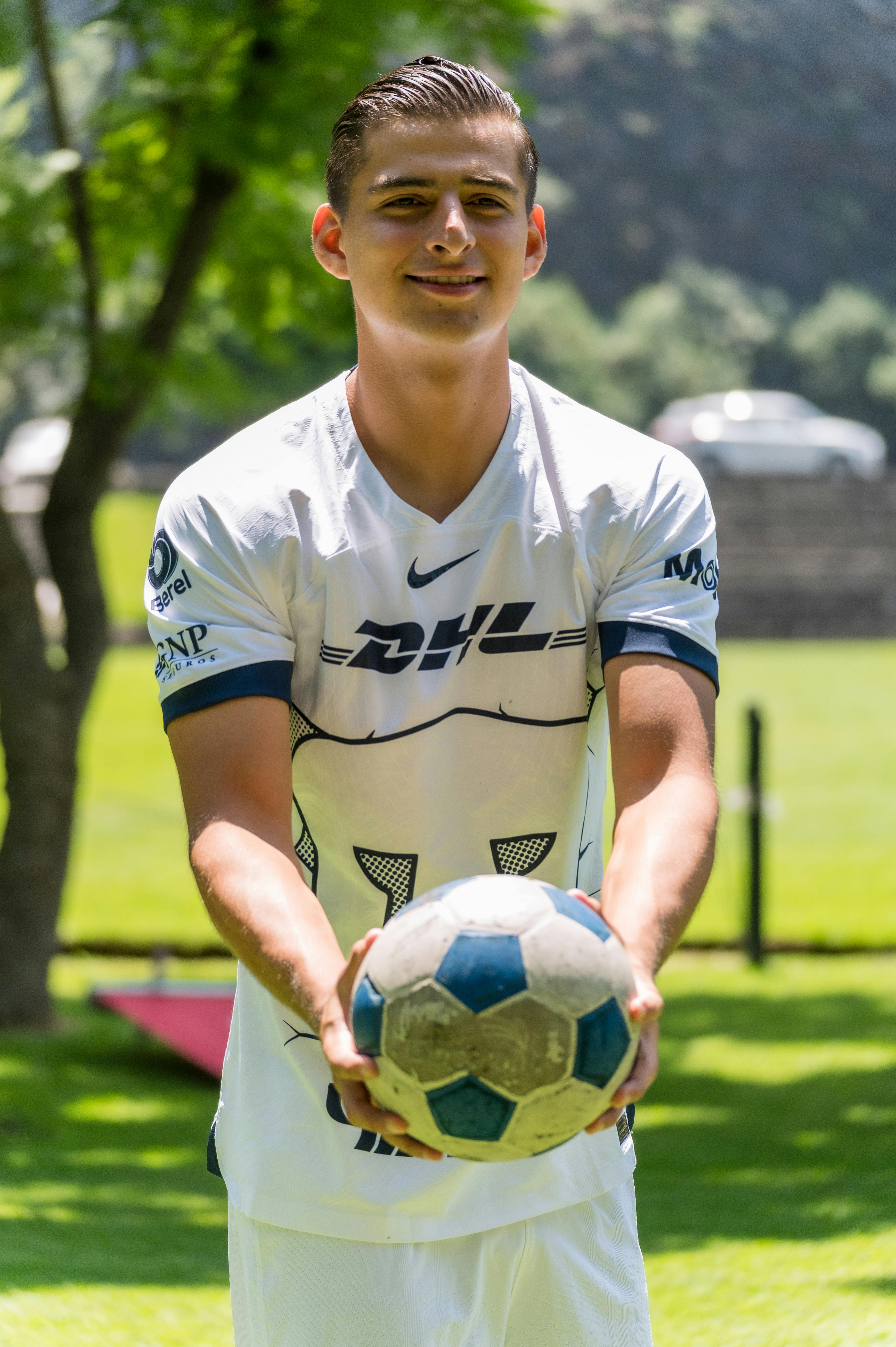 Smiling young male in soccer jersey holding a soccer ball outdoors in a sunny park.