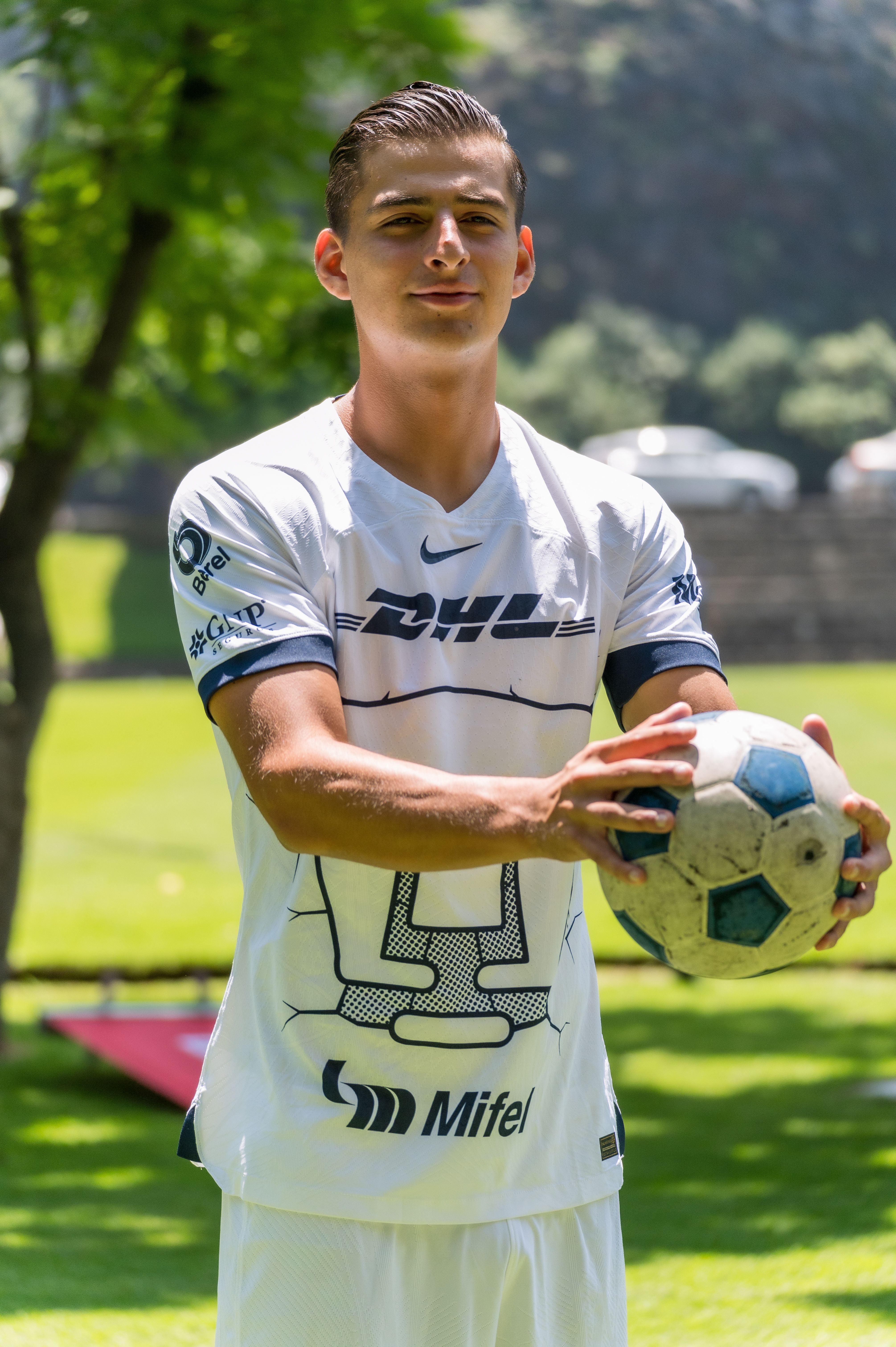 A young male soccer player in sportswear holding a ball in a sunny outdoor setting.