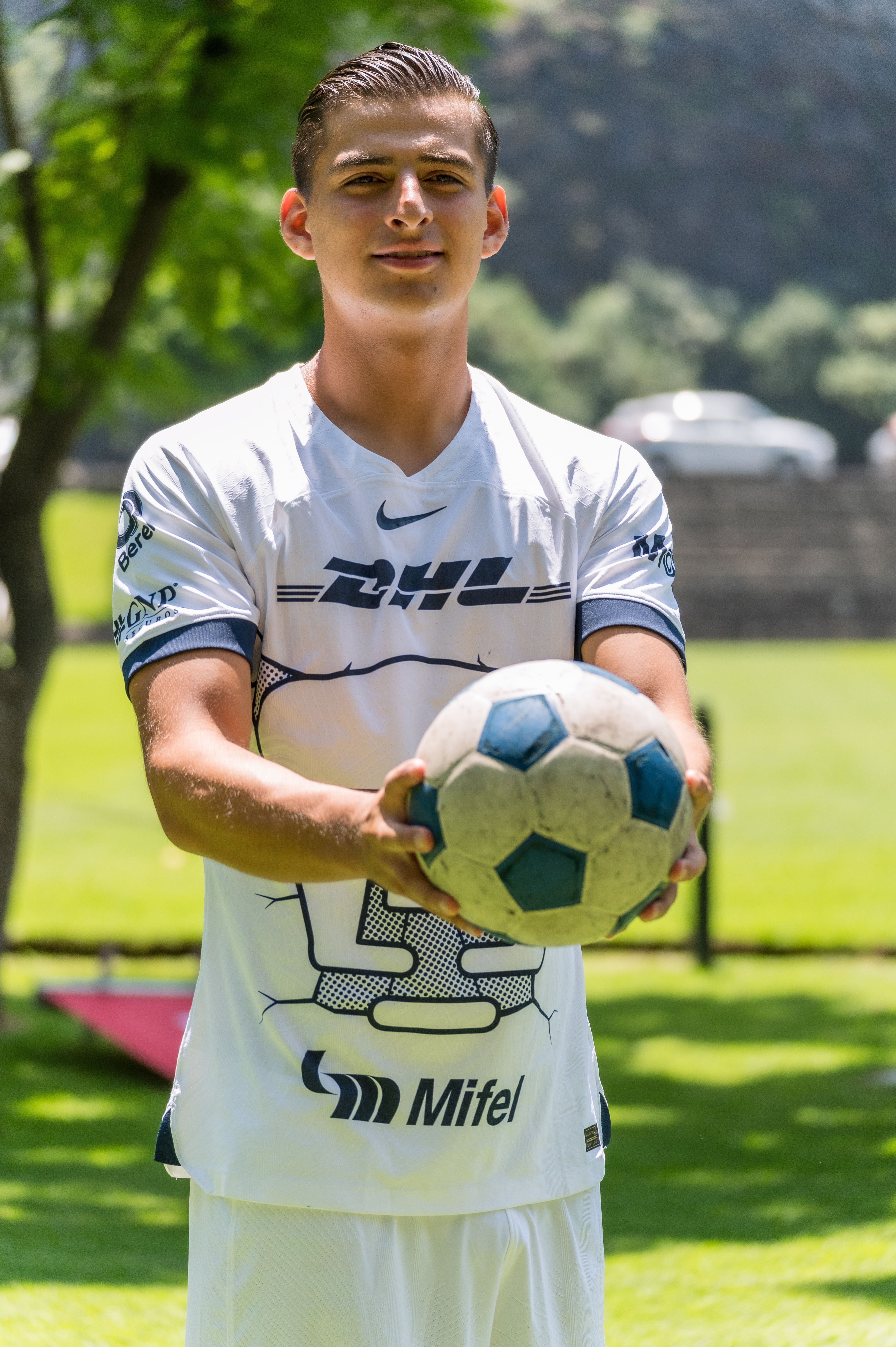 A young male football player holding a soccer ball on a sunny day outdoors in a park.