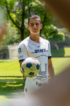 Teenage boy in sports jersey holding a soccer ball on a sunny day outdoors.