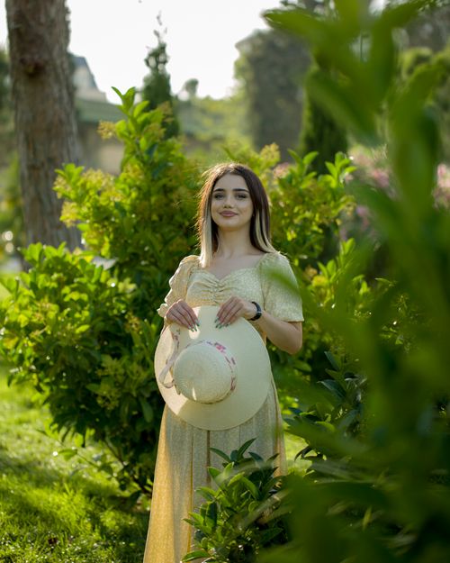 Free A woman in a green sundress stands in a lush garden holding a straw hat, surrounded by vibrant foliage. Stock Photo