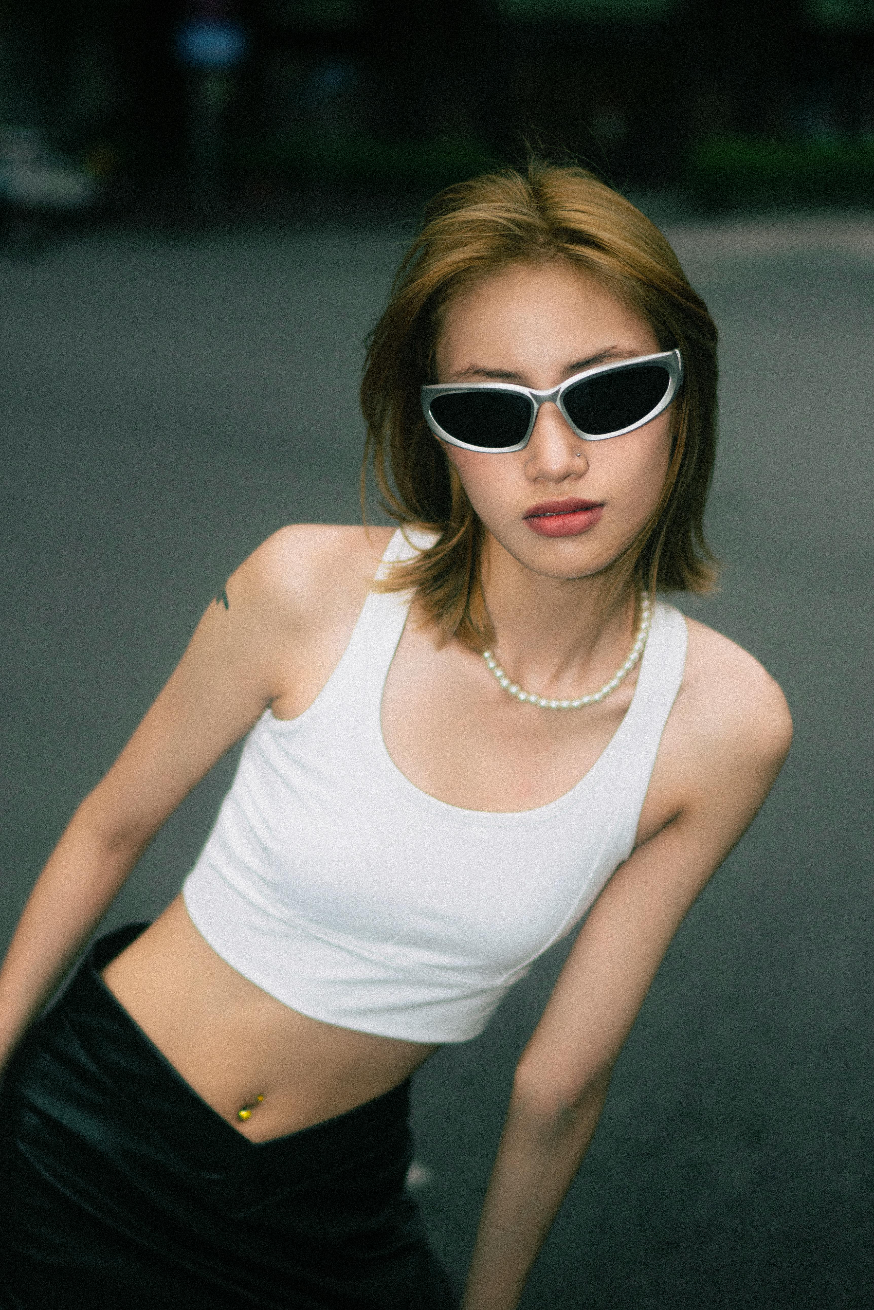 Fashionable woman in white tank top posing outdoors with sunglasses.