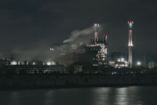 Night view of a large industrial factory with chimneys emitting smoke by the river in Ahmedabad, India.