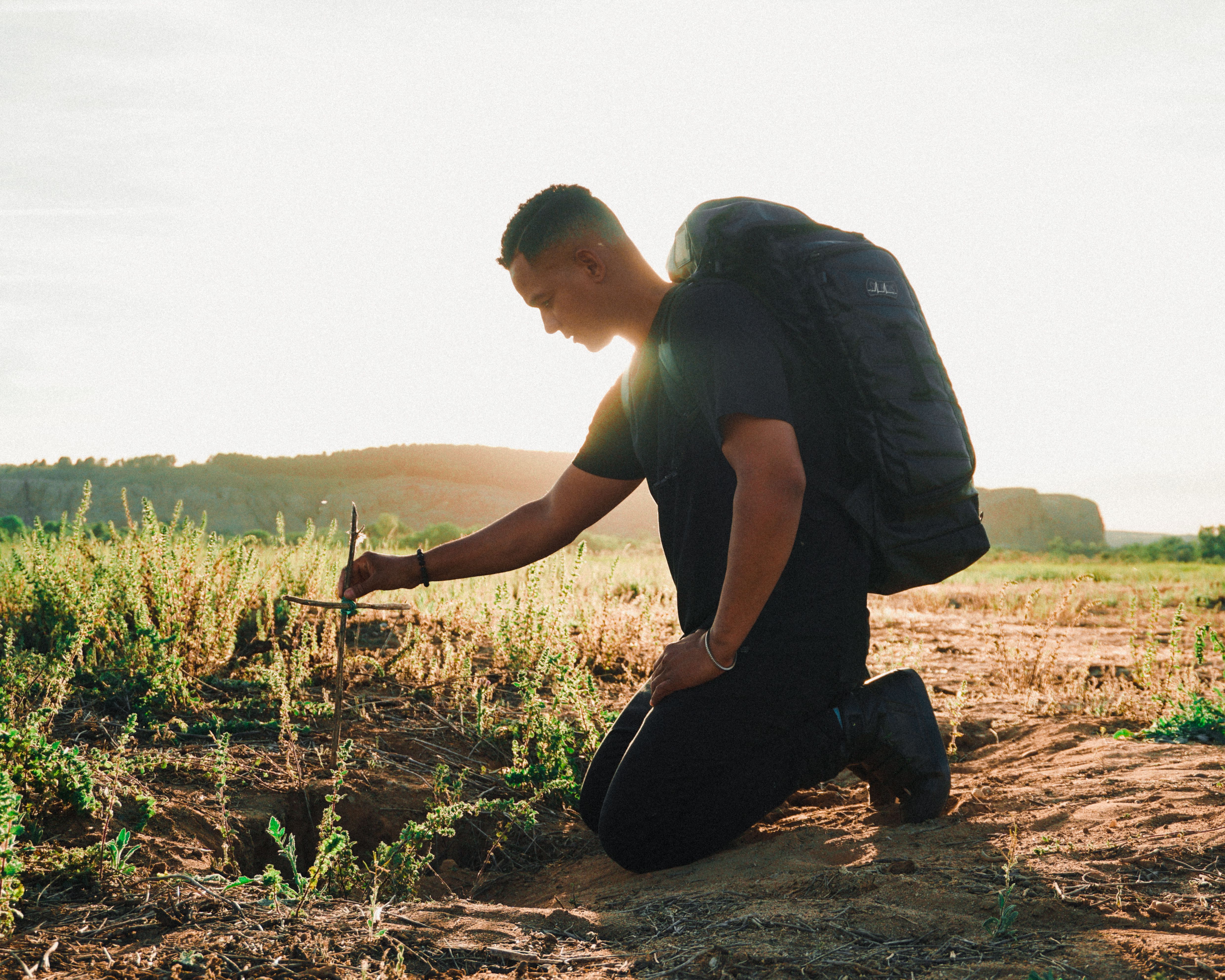 Traveler kneeling at a rural field cross, backlit by sunlight, symbolizing reflection and solitude.