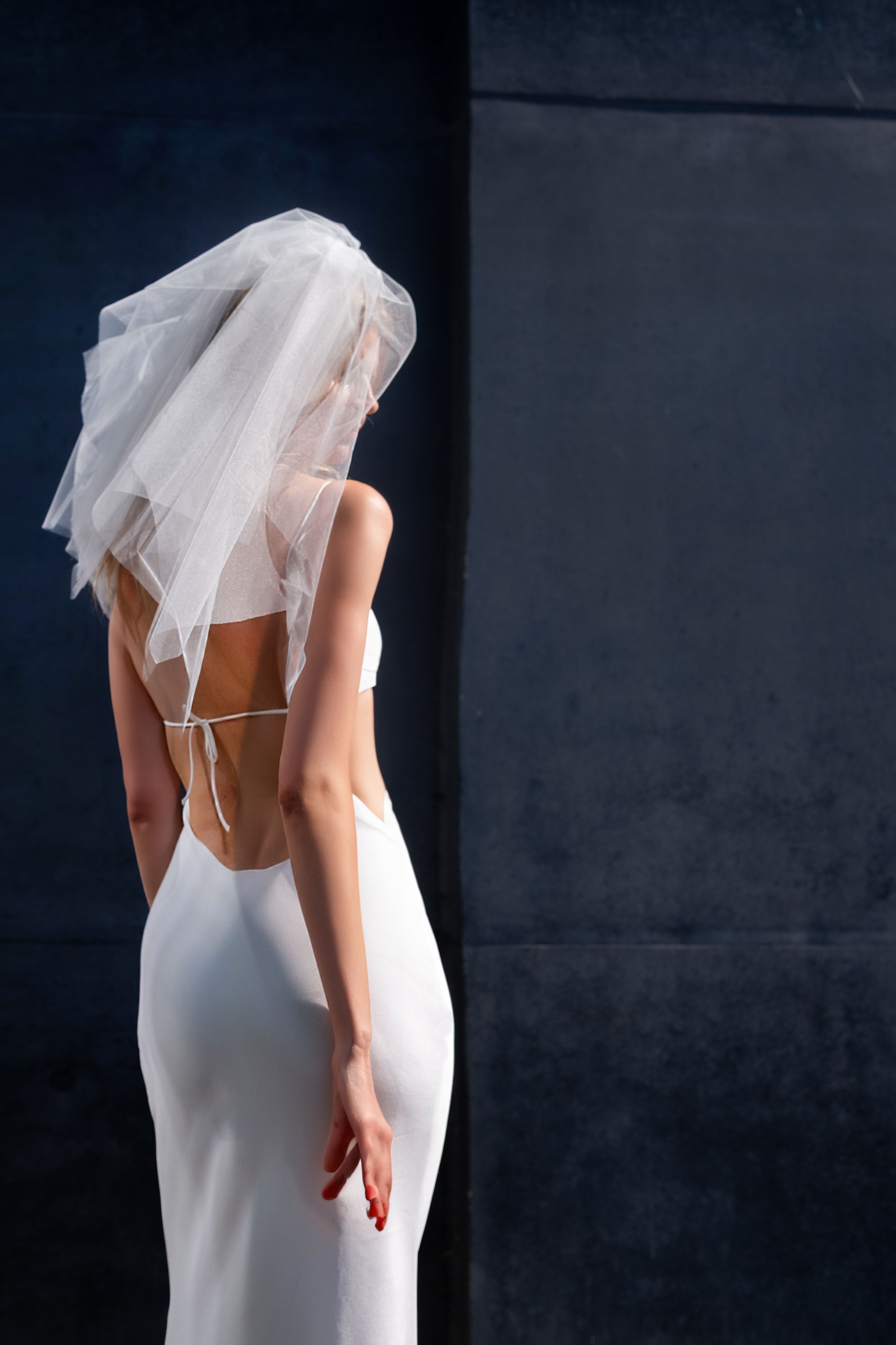 A fashion-forward bride stands elegantly in a white dress with veil, set against a minimalist dark wall.