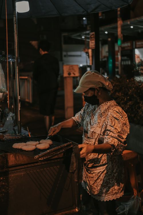 Free Masked street vendor grills bread at night, showcasing urban culinary traditions. Stock Photo