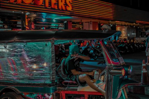 A vibrant tuk-tuk and a person relaxing in neon-lit urban setting at night.