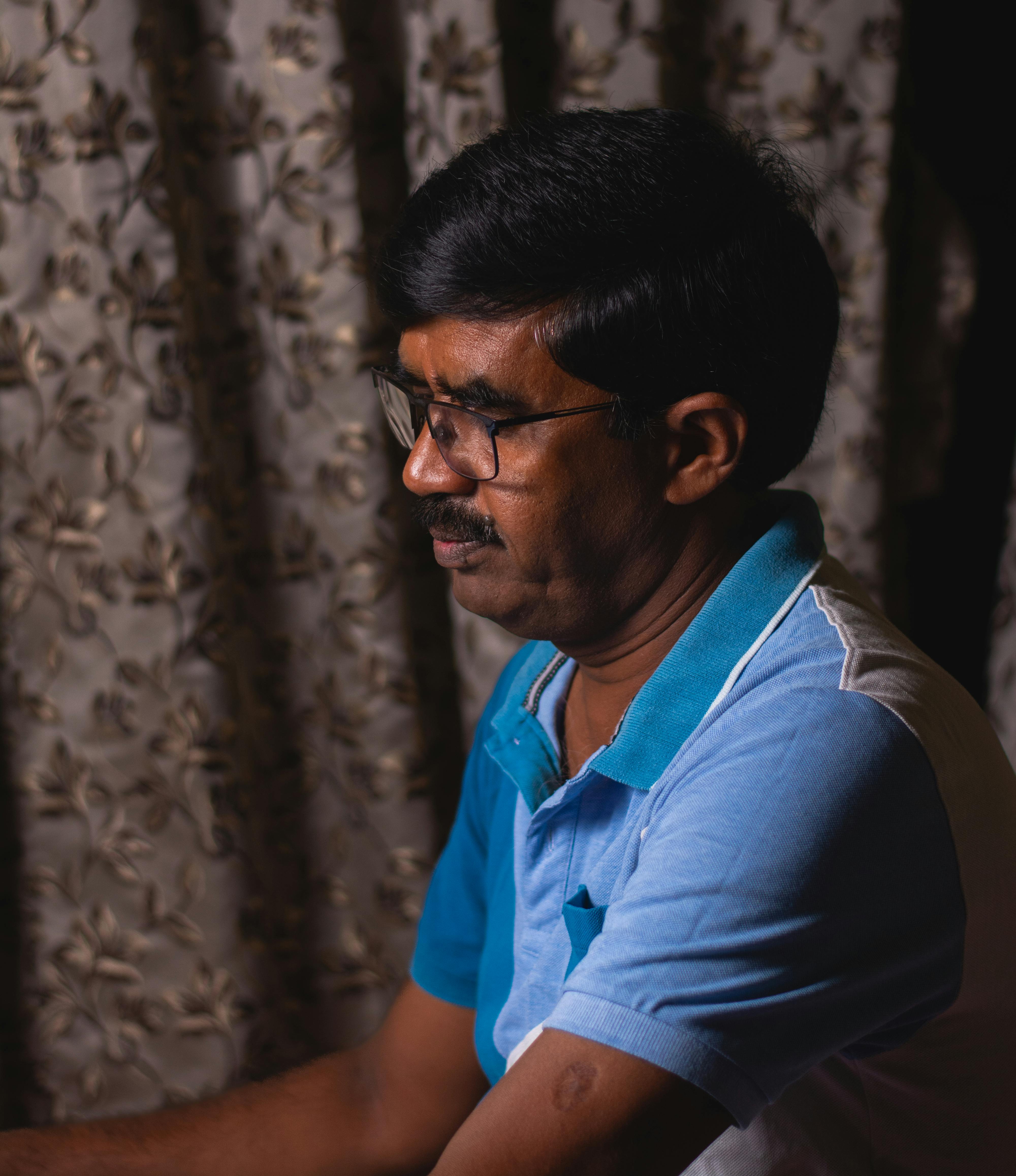 Portrait of an adult man with black hair and glasses, looking contemplative indoors.
