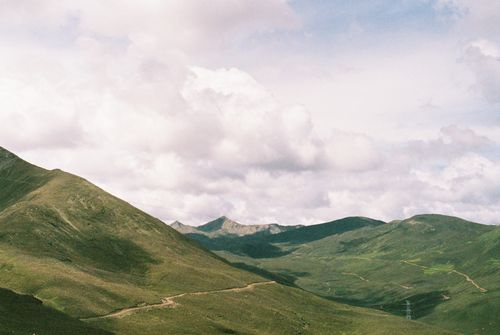 Free Explore scenic green hills with a winding path and mountainous backdrop under a vivid sky. Stock Photo