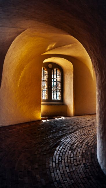 Artistic view of a curved brick corridor leading to a stained glass window illuminated by natural light.
