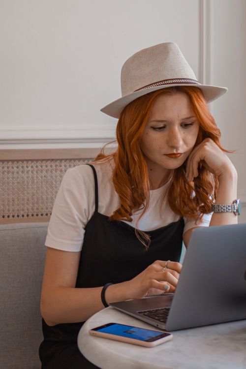 Free Redhead woman wearing a hat, focused on laptop work with a smartphone on the table. Stock Photo