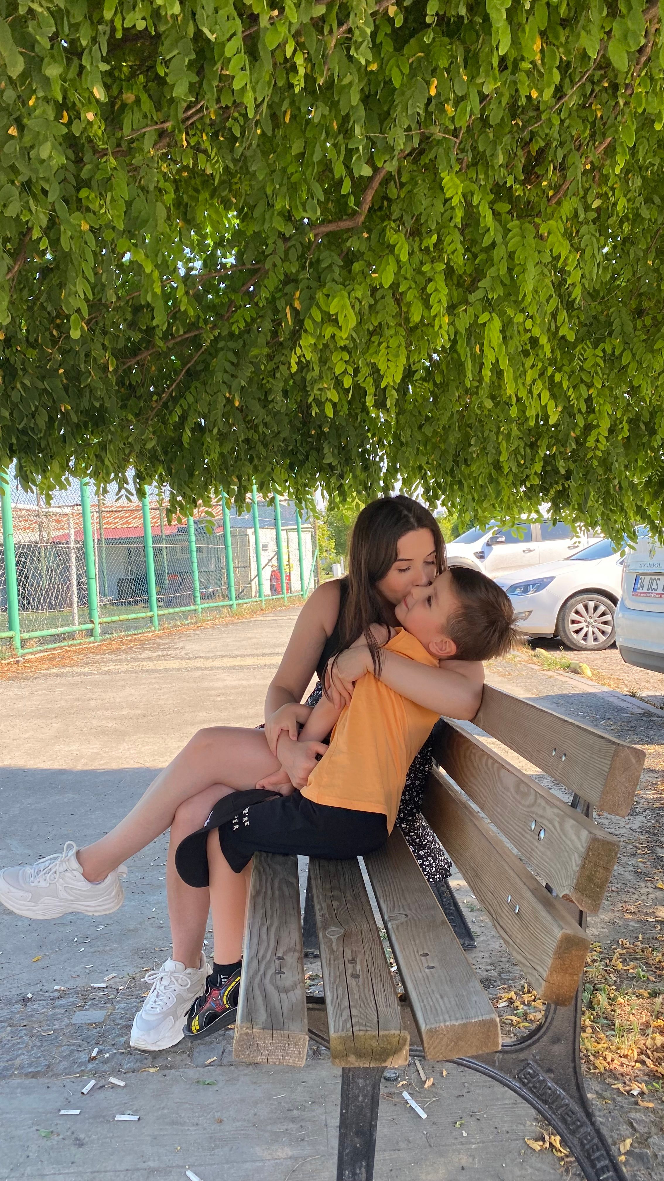 Woman and young child embracing on bench under a tree in an urban park setting.