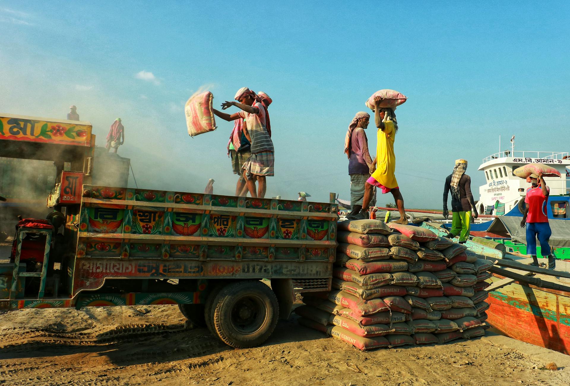 Workers unloading bags from a colorful truck by the waterfront, showcasing teamwork and labor.
