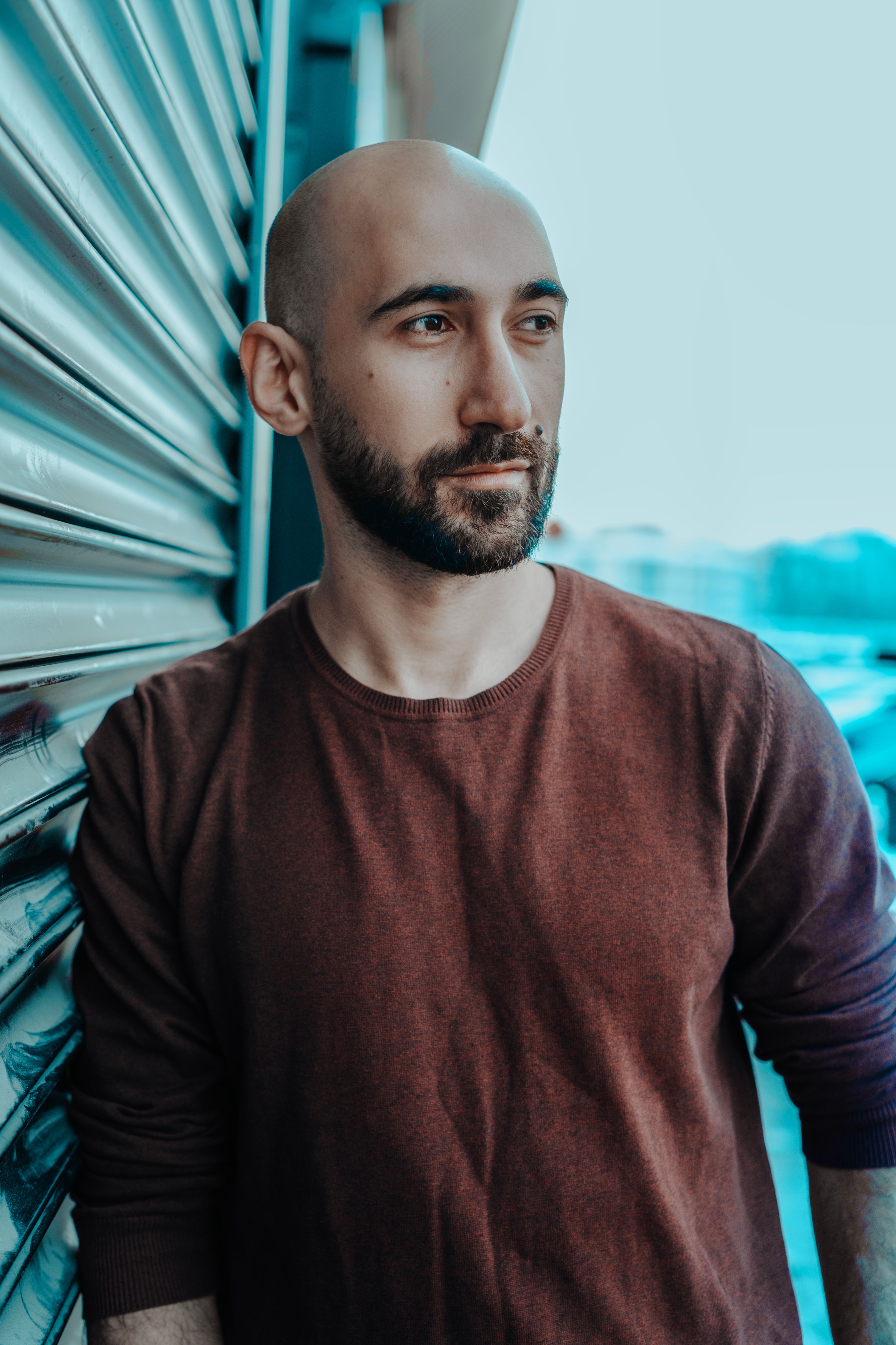 Close-up portrait of a bald man in a maroon sweatshirt leaning against a metal surface.