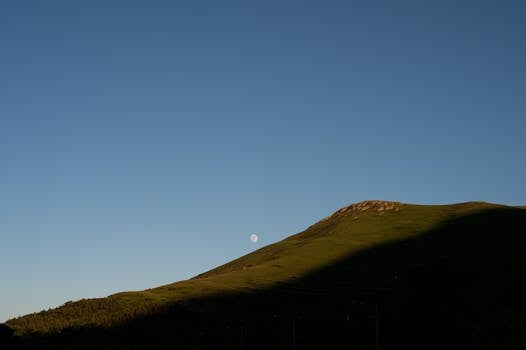 Serene mountain landscape under a clear sky with the moon rising, captured at dusk.
