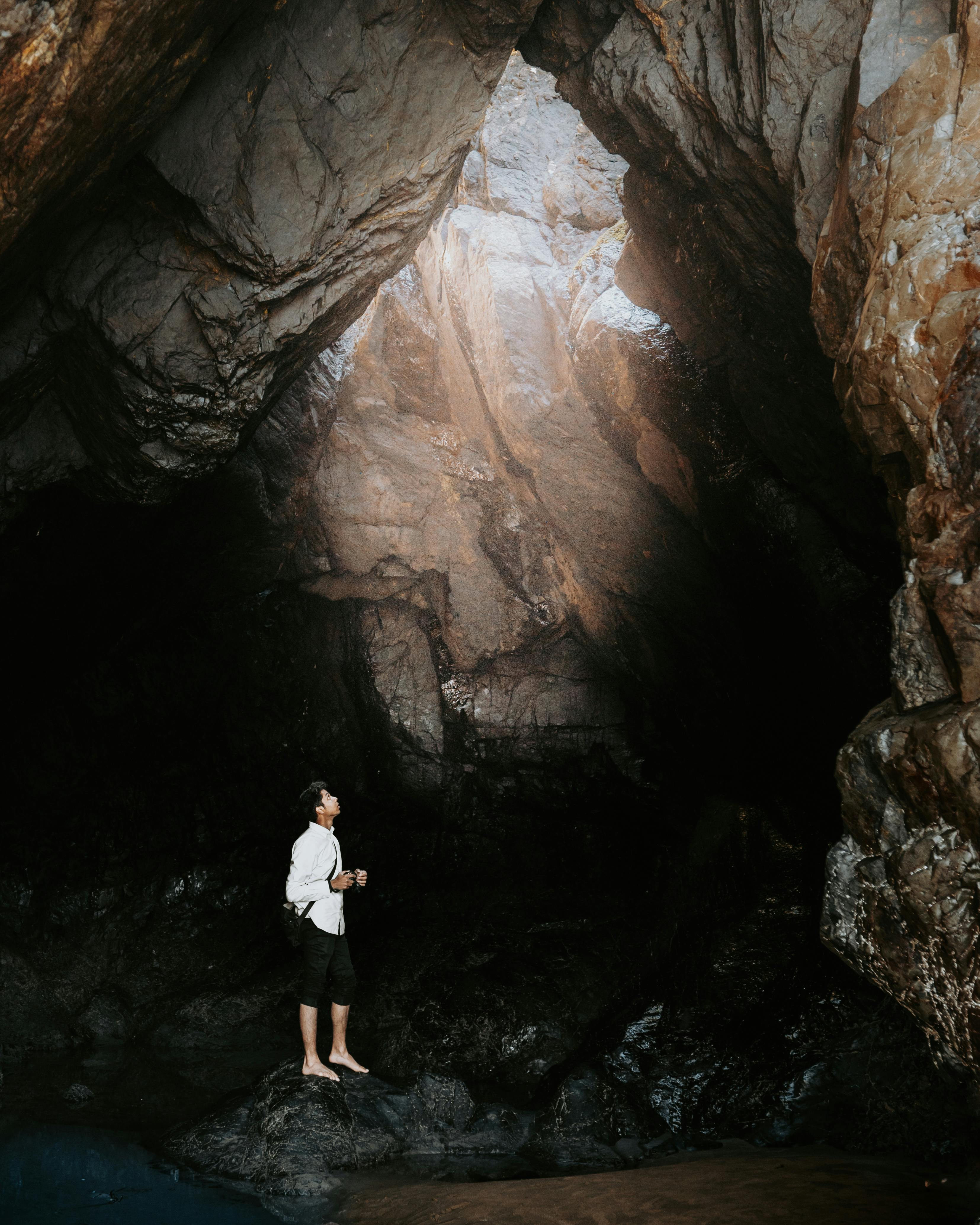 A man in a white shirt explores a large rock cave with dramatic lighting from above.