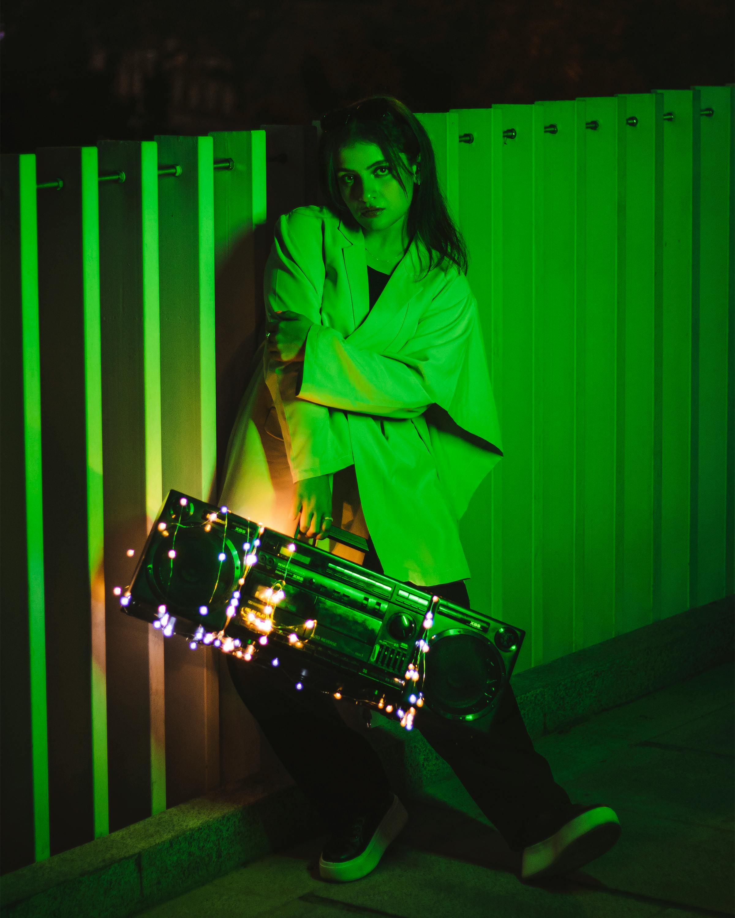 A woman stands with a lit boombox against a green-lit backdrop, creating a dramatic and moody scene.