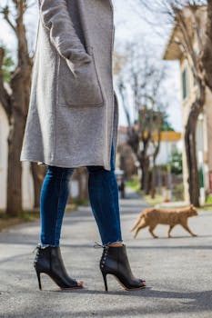 A fashionable woman in a gray coat and open-toe heels walking on a city street with a cat nearby.